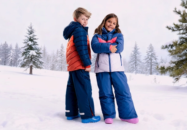 Zwei Kinder in bunten Winterkleidern stehen in einer verschneiten Landschaft, umgeben von hohen immergrünen Bäumen und einer nebligen Atmosphäre.