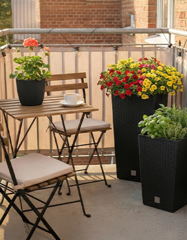 Gemütlicher Balkon mit Holztisch und Stühlen, einer Tasse und Pflanzkästen voller leuchtend roter und gelber Blumen sowie grüner Kräuter.