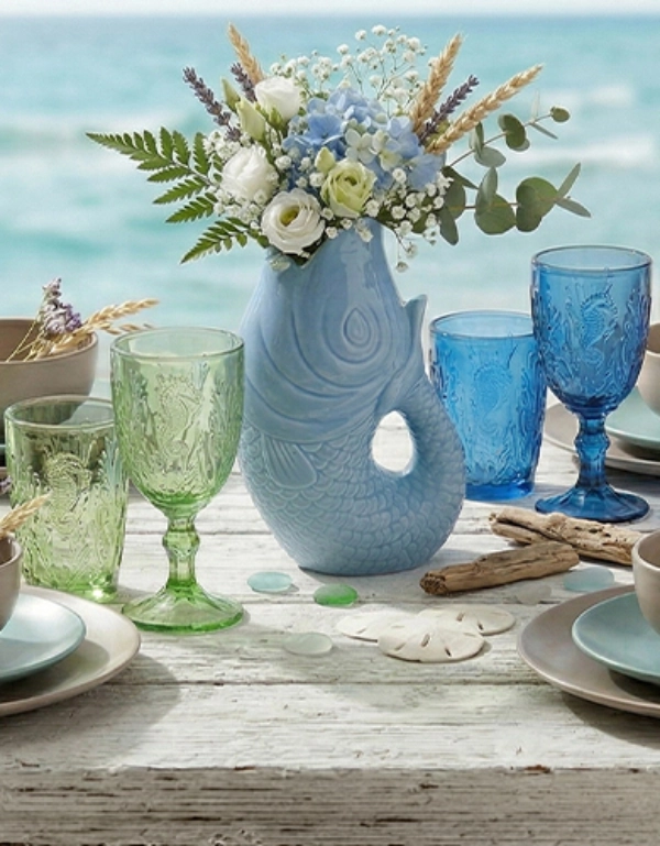 Maritim gedeckter Tisch mit farbigen Gläsern, Geschirr und dekorativer Vase mit Blumen vor Meereskulisse – stilvolle Tischdekoration für sommerliche Wohlfühlmomente