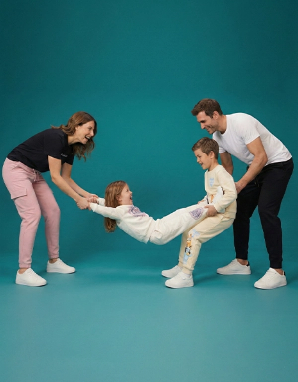 Eine Familie interagiert spielerisch miteinander und hält ein Kind zwischen zwei Erwachsenen; alle sind lässig gekleidet vor einem türkisfarbenen Hintergrund.