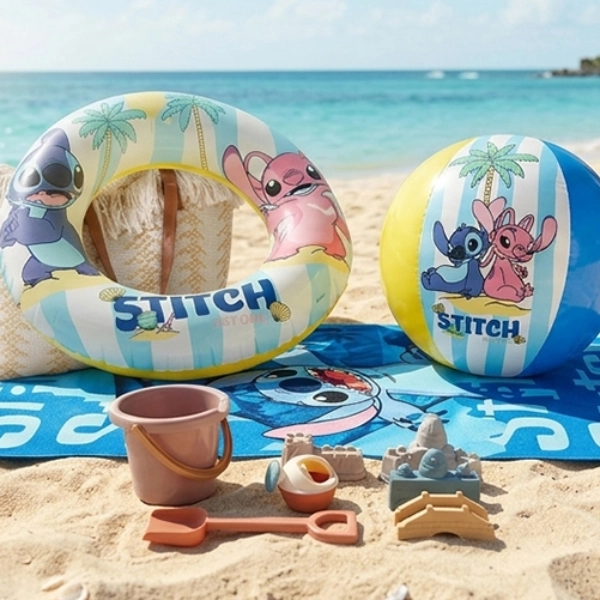 Strandtuch, Schwimmring, Ball und Sandspielzeug mit Lilo‑&‑Stitch‑Motiv liegen am Sandstrand neben türkisfarbenem Meer.