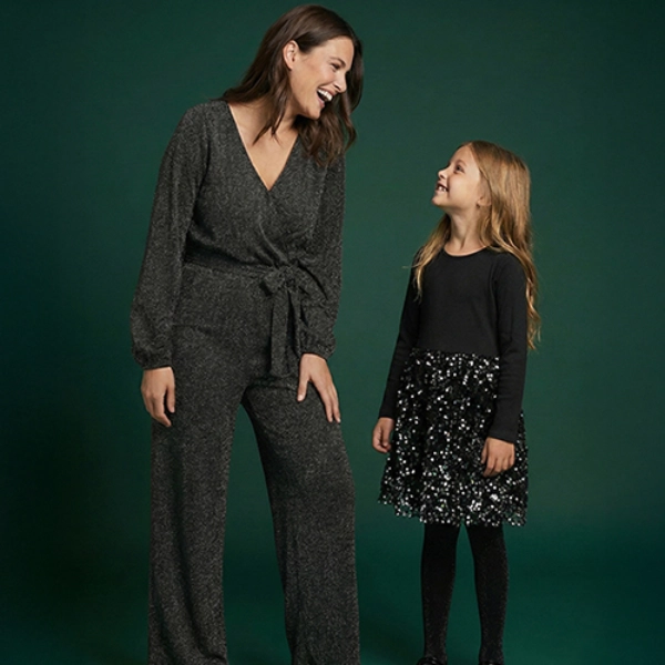 Eine Frau in einem glitzernden schwarzen Overall steht neben einem Mädchen in einem schwarzen Oberteil mit einem Paillettenrock, beide lächeln vor einem dunkelgrünen Hintergrund.