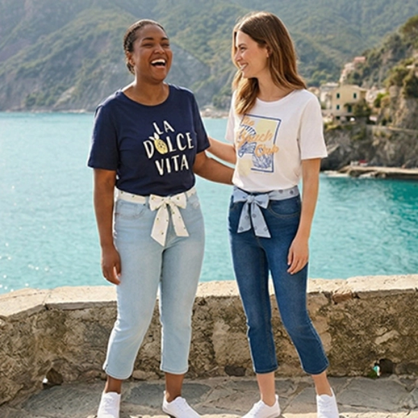 Zwei Frauen stehen an einer Steinterrasse am Meer, im Hintergrund farbenfrohe Häuser an einer Klippe an einem sonnigen Tag.