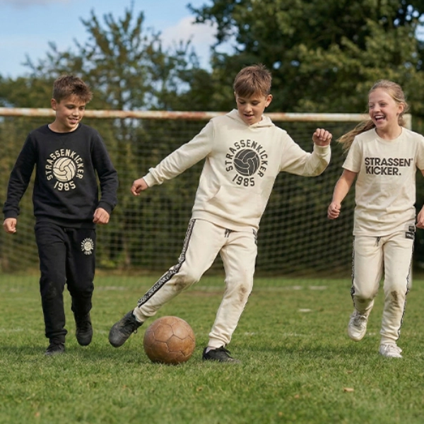 Drei Kinder in lässiger Sportkleidung spielen Fußball auf einer Grasfläche, mit einem Tor im Hintergrund.
