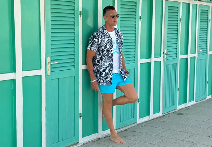 Ein Mann in einem tropischen Hemd und blauen Shorts lehnt lässig an grünen Strandhütten und genießt den sonnigen Tag.