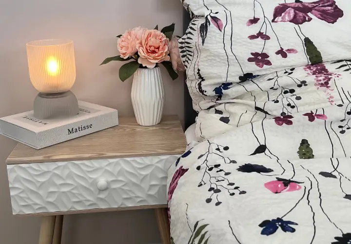 Eine gemütliche Nachttischszene mit einer leuchtenden Lampe, einer Blumenvase mit rosa Rosen und einer gemusterten Bettdecke vor einer sanften, neutralen Wand.