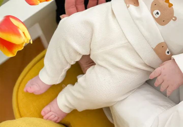 Eine Nahaufnahme der Beine eines Babys in einem Schlafanzug mit Bärenmuster, das auf einem Schoß liegt, mit leuchtenden Tulpen im Vordergrund. Eine Nahaufnahme der Beine eines Babys in einem Schlafanzug mit Bärenmuster, das auf einem Schoß liegt, mit leuchtenden Tulpen im Vordergrund.