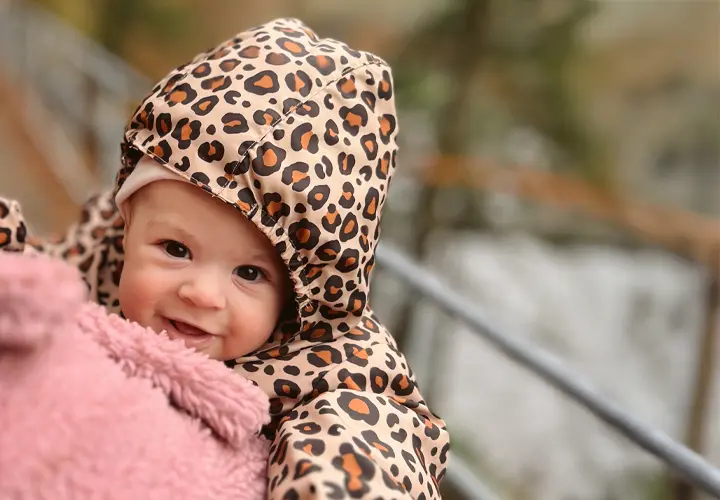 Ein Baby in einer Kapuzenjacke mit Leopardenmuster kuschelt sich in eine weiche rosafarbene Decke vor einer natürlichen Kulisse. Ein Baby in einer Kapuzenjacke mit Leopardenmuster kuschelt sich in eine weiche rosafarbene Decke vor einer natürlichen Kulisse.