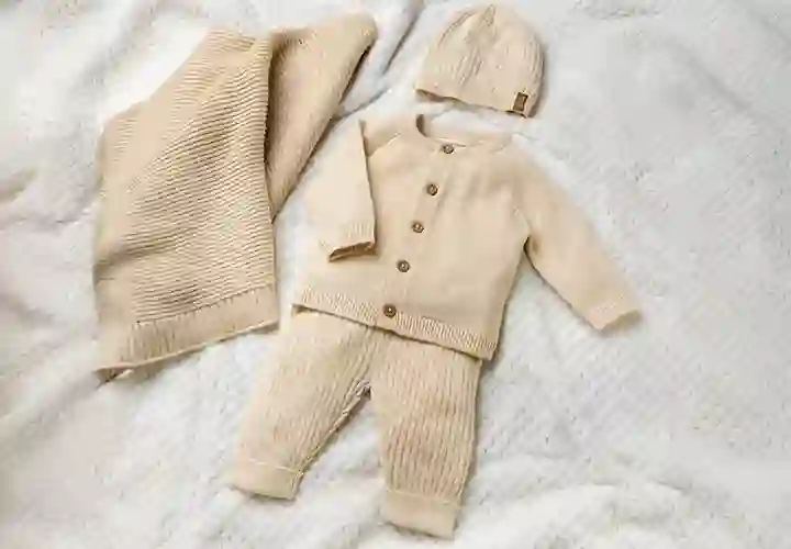Ein weiches Babyoutfit in Beige mit Strickjacke, Hose und passender Mütze, präsentiert auf einer strukturierten Decke.