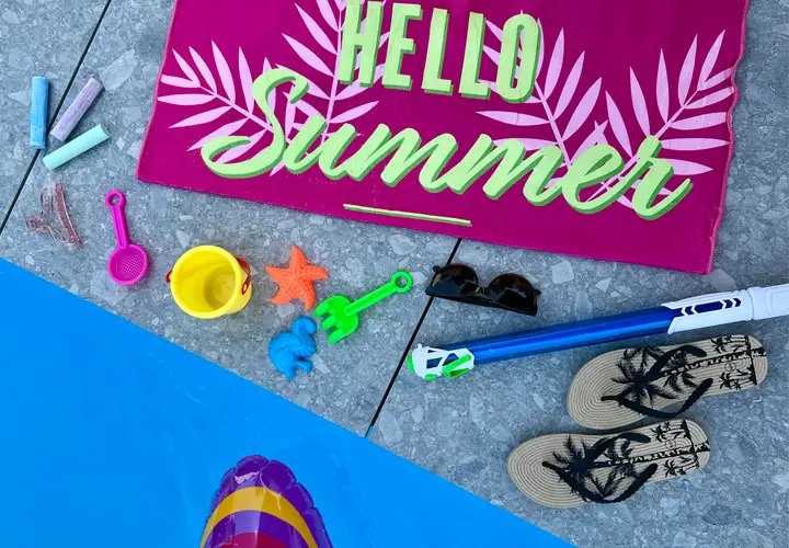 Eine lebhafte Sommerszene mit einem rosa Handtuch mit der Aufschrift „Hello Summer“, Strandspielzeug, Sonnenbrillen und Flip-Flops neben einem Pool.