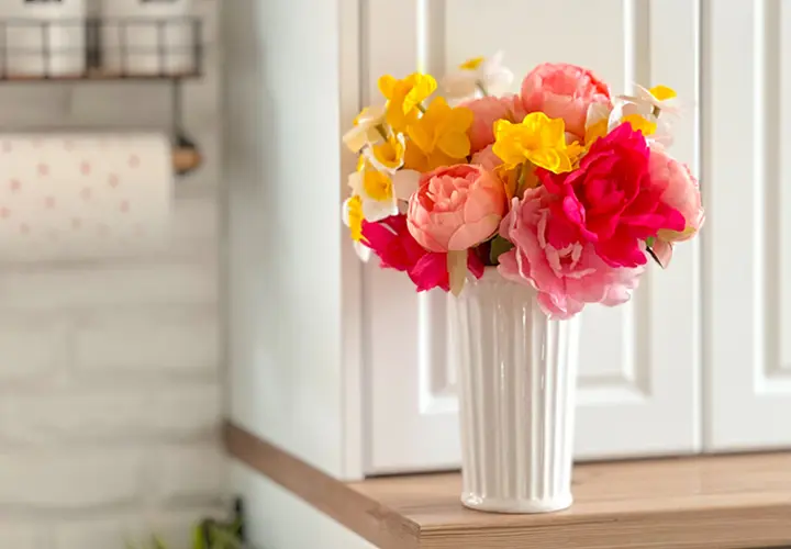 Bunte Kunstblumen in einer weißen Vase, ausgestellt auf einem Holzregal vor einem hellen Küchenhintergrund.