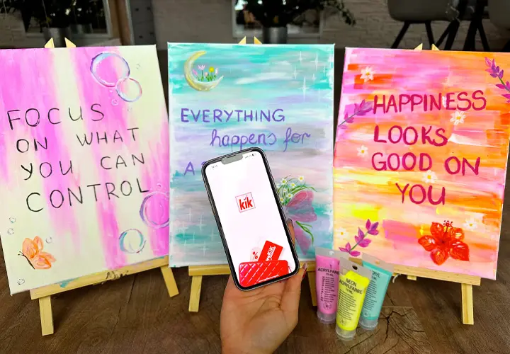 Eine Hand hält ein Smartphone mit der KiK-App, daneben stehen drei bunte Gemälde mit motivierenden Zitaten auf Staffeleien.