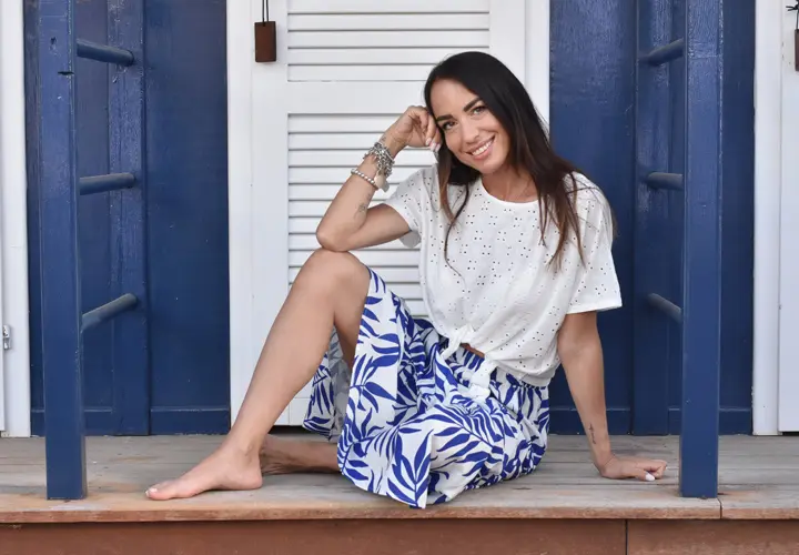 Eine Frau sitzt anmutig auf einer Veranda, trägt ein weißes Oberteil und einen blauen, geblümten Rock, die Hand auf dem Kinn, tief in Gedanken versunken.