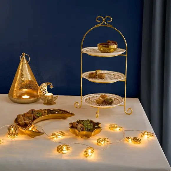 Ein wunderschön arrangierter Desserttisch mit einem goldenen dreistöckigen Ständer mit Gebäck, Laternen und funkelnden Lichterketten vor einem tiefblauen Hintergrund.