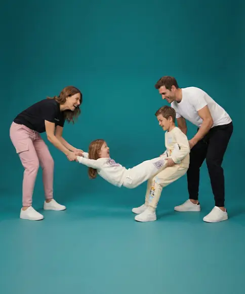 Eine Familie interagiert spielerisch miteinander und hält ein Kind zwischen zwei Erwachsenen; alle sind lässig gekleidet vor einem türkisfarbenen Hintergrund.