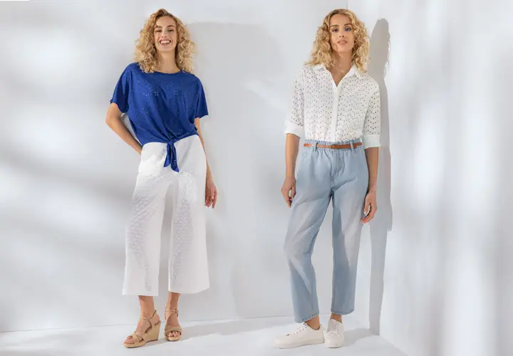 Zwei Frauen modellieren stilvolle Outfits: die eine in einem blauen Oberteil und einer weißen Culottes, die andere in einer weißen Bluse und einer hellblauen Hose vor einem hellen Hintergrund.