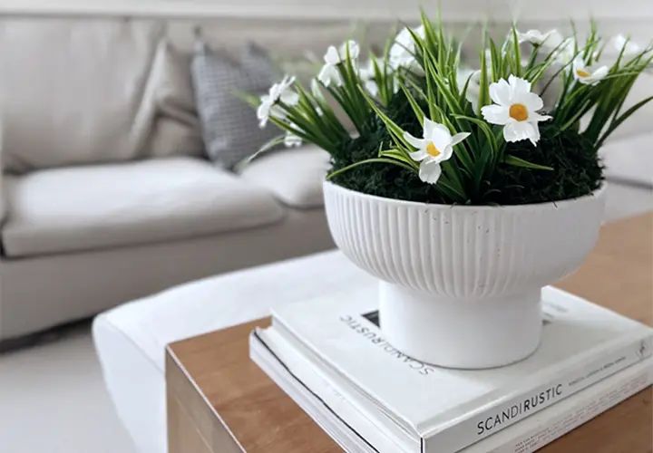 Eine weiße Schale mit künstlichen Gänseblümchen und Grün steht auf gestapelten Büchern auf einem Holzcouchtisch, im Hintergrund ein helles Sofa.