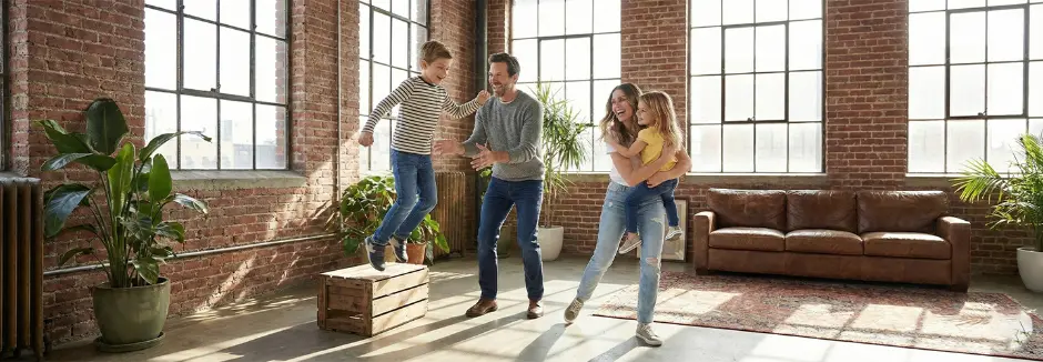 Eine fröhliche Familie spielt gemeinsam in einem großzügigen, lichtdurchfluteten Raum mit Backsteinwänden und Pflanzen und genießt ausgelassene Momente voller Spaß und Verbundenheit.
