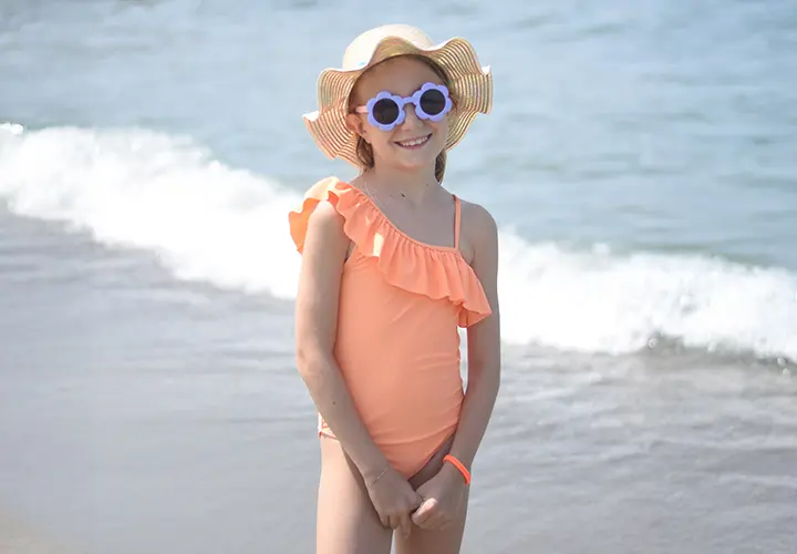 Ein Kind im korallfarbenen One-Shoulder-Badeanzug mit Rüschen steht am Strand und trägt einen breitkrempigen Strohhut