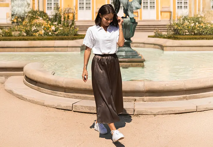Eine Frau in weißem Shirt und braunem Rock geht an einem Brunnen entlang, umgeben von Grünflächen, mit gelben Gebäuden im Hintergrund.