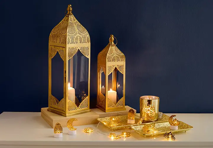 Zwei aufwendig gestaltete goldene Laternen mit Kerzen, ein Ziertablett und festliche Lichter vor einer tiefblauen Wand.