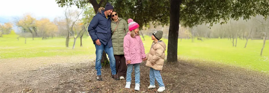 Eine Familie steht gemeinsam in einem grünen Park, warm gekleidet für kühles Wetter, umgeben von Bäumen und einer ruhigen Landschaft.