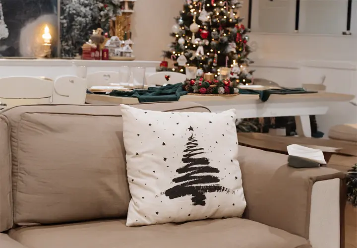 Ein gemütliches Wohnzimmer mit einem Zierkissen mit Weihnachtsbaum-Motiv, festlicher Dekoration und einem schön beleuchteten Weihnachtsbaum.