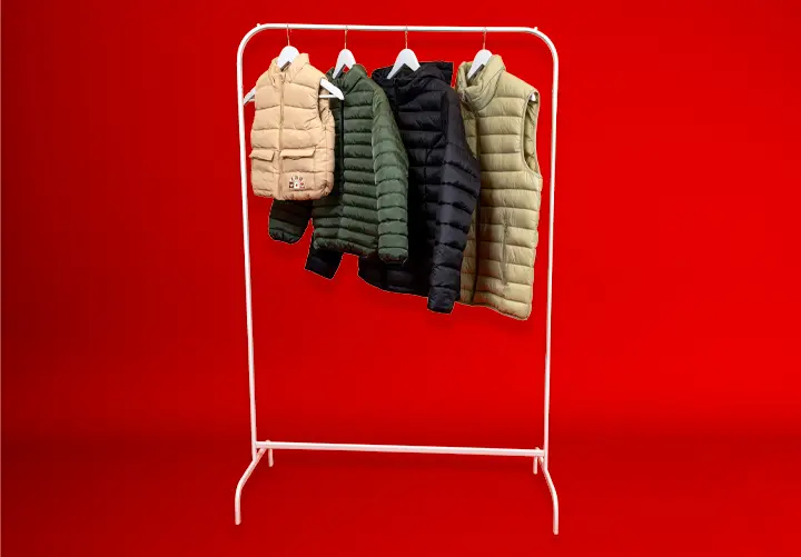 Ein weißer Kleiderständer zeigt vier Steppwesten in verschiedenen Farben – Beige, Dunkelgrün, Marineblau und Oliv – vor rotem Hintergrund. Ein weißer Kleiderständer zeigt vier Steppwesten in verschiedenen Farben – Beige, Dunkelgrün, Marineblau und Oliv – vor rotem Hintergrund.