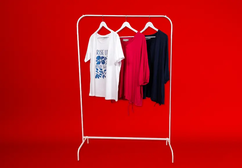 Drei stilvolle Oberteile hängen auf einem weißen Kleiderständer vor einem leuchtend roten Hintergrund: ein weißes T-Shirt mit Aufdruck, eine rosa Bluse und ein marineblaues Hemd.