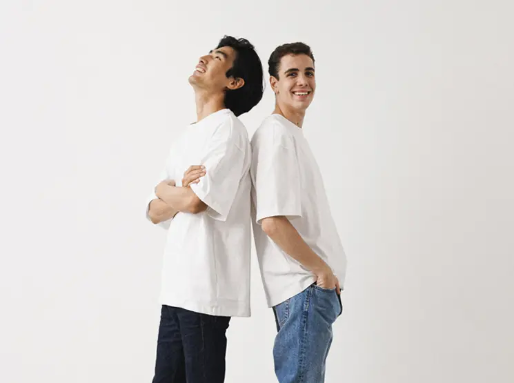Basic Herrenmode Zwei junge Männer in weißen T-Shirts stehen Rücken an Rücken vor einem schlichten weißen Hintergrund und lächeln entspannt.