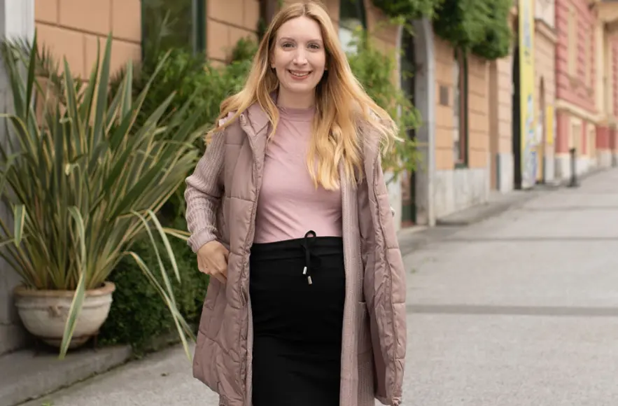 Lagenlook, Layering oder Zwiebellook  Eine Frau steht auf einem Gehweg, trägt einen leichten Steppmantel über einem rosa Oberteil und einem schwarzen Rock, umgeben von üppigem Grün.