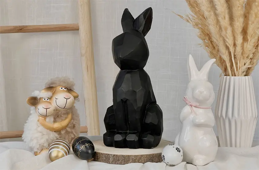 Eine schwarze geometrische Hasenfigur steht zwischen einer Schaffigur und einem weißen Hasen, umgeben von Dekoeiern und getrocknetem Gras.