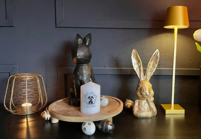 Ein österliches Holz-Arrangement mit zwei Hasendekorationen, einer Kerze und Dekoeiern auf einem runden Tablett vor dunklem Hintergrund.