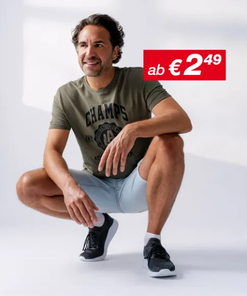 Ein Mann hockt in lässiger Pose, trägt ein grünes T-Shirt und hellblaue Shorts, gepaart mit schwarzen Sportschuhen, vor einem weichen Hintergrund.