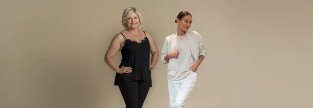 Zwei Frauen stehen nebeneinander, eine in einem schwarzen Oberteil und Leggings, die andere in einer hellen Jacke und Hose, vor einem neutralen Hintergrund.