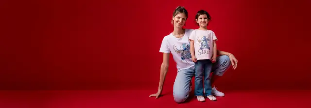 Eine Frau und ein Kind posieren vor einem leuchtend roten Hintergrund und tragen passende Lilo & Stitch T-Shirts und lässige Jeans