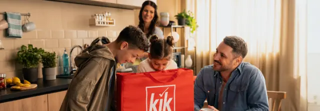 Eine Familie packt freudig eine rote kik-Liefertasche in einer hellen Küche aus, die mit Pflanzen und fröhlicher Dekoration gefüllt ist.