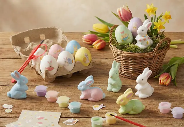 Farbenfrohe Osterdekorationen mit bemalten Eiern, pastellfarbenen Hasen und Malutensilien auf einem rustikalen Holztisch mit Blumen.
