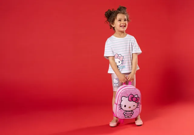 Ein Kind steht vor einem roten Hintergrund, trägt ein gestreiftes Hello Kitty-Shirt und Shorts und hält einen rosa Hello Kitty-Rucksack in der Hand