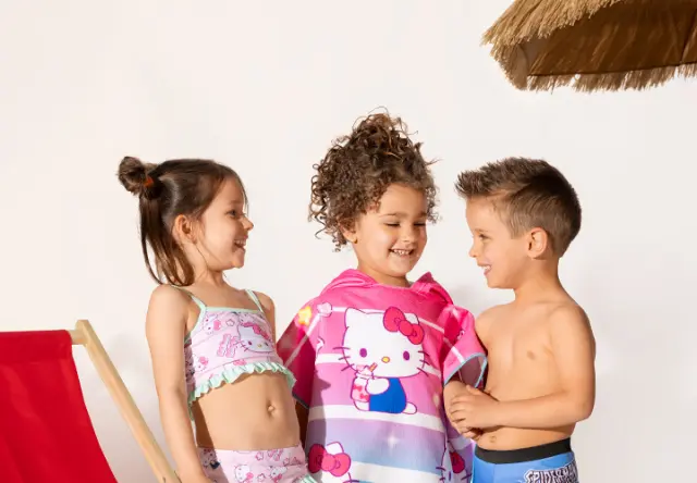 Drei Kinder in Badekleidung stehen zusammen, eines davon in ein buntes Hello Kitty-Handtuch gewickelt, im Hintergrund ein Strandstuhl und ein Strohsonnenschirm.