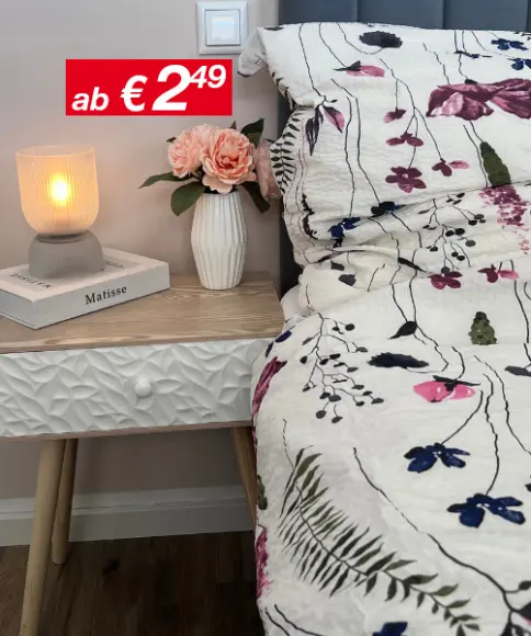 Eine gemütliche Nachttischszene mit einer leuchtenden Lampe, einer Blumenvase mit rosa Rosen und einer gemusterten Bettdecke vor einer sanften, neutralen Wand.