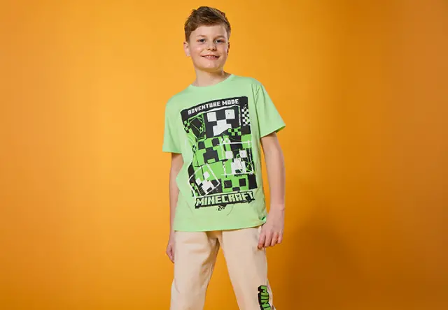 Eine Person posiert vor einem leuchtend orangefarbenen Hintergrund und trägt ein grünes Minecraft-T-Shirt und eine hellbeige Hose.