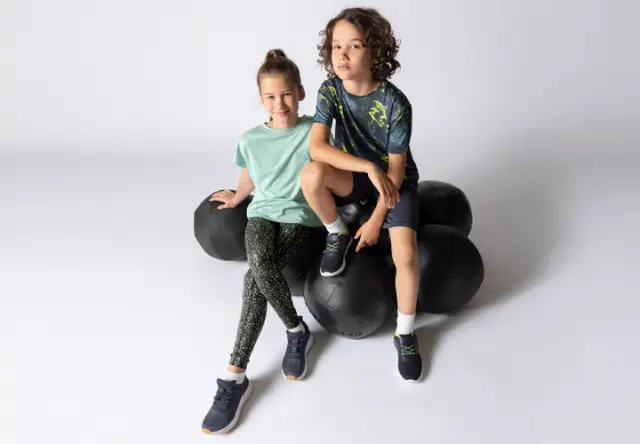 Zwei Kinder sitzen auf einer Gruppe schwarzer Gymnastikbälle, gekleidet in bunte Sportbekleidung und Sportschuhe, vor neutralem Hintergrund.