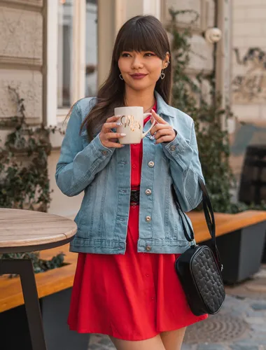 Eine Frau in Jeansjacke und rotem Kleid hält einen Kaffeebecher, während sie im Freien neben einem Tisch und Holzfässern steht.