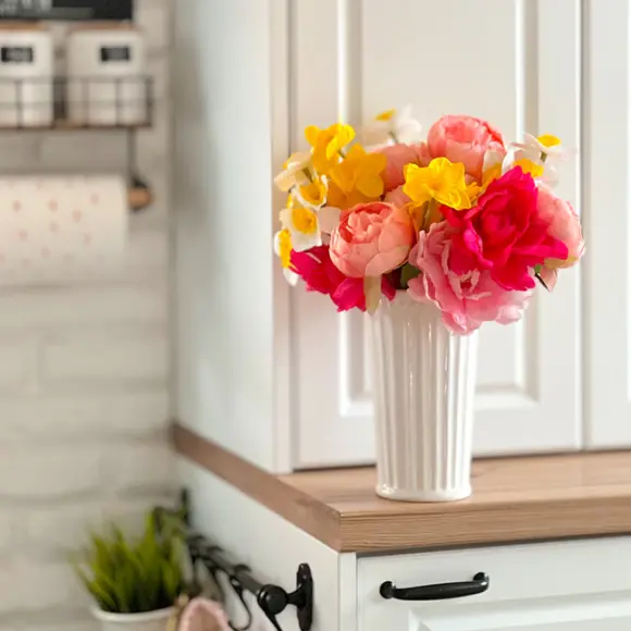 Bunte Kunstblumen in einer weißen Vase, ausgestellt auf einem Holzregal vor einem hellen Küchenhintergrund.