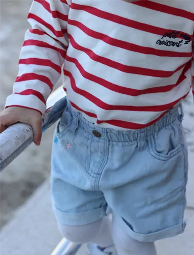Ein Kind, das ein rot-weiß gestreiftes langärmeliges Hemd und hellblaue Jeans-Shorts trägt, hält sich an einem Geländer fest.
