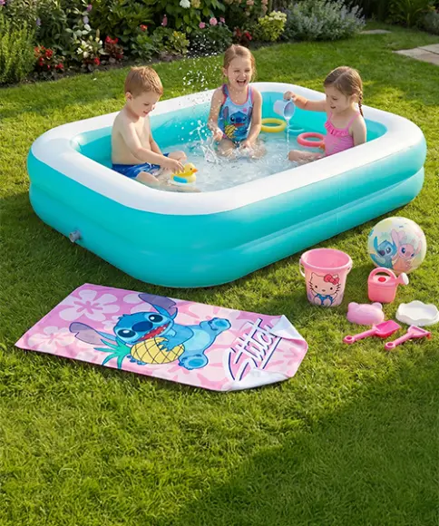 Drei Kinder spielen in einem kleinen aufblasbaren Pool auf einer Wiese, daneben liegen Spielzeuge, ein Strandtuch und ein Ball in einem sonnigen Garten.