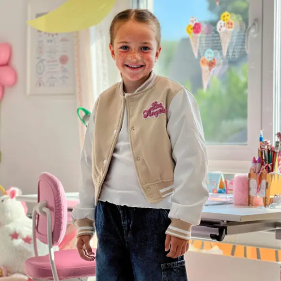 Ein Kind in einer stylischen beige-weißen Collegejacke steht in einem bunten, fröhlichen Raum voller Bastelmaterialien und Dekorationen.