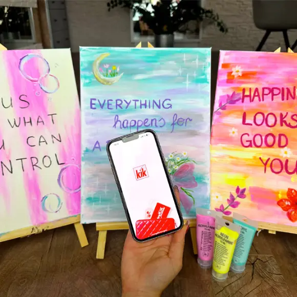 Eine Hand hält ein Smartphone mit der KiK-App, daneben stehen drei bunte Gemälde mit motivierenden Zitaten auf Staffeleien.