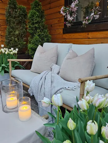 Gemütliche Sitzgelegenheiten im Freien mit weichen Kissen, einer gestrickten Decke, leuchtenden Kerzen und blühenden Tulpen vor einem Holzhintergrund.